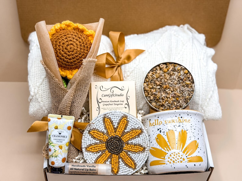 May include: A gift box containing a crocheted sunflower in burlap, a white cable-knit blanket with a gold ribbon, a CareGiftStudio soap, a tin of dried chamomile, a beaded sunflower coaster, a chamomile hand cream, and a hello sunshine mug.