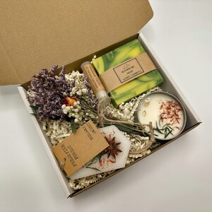 May include: A gift box filled with a green bar of soap labelled "Handmade Soap Lemon Verbena", a small white candle with red and yellow flower petals, a small tube of lip balm, and a small tag with the text "Fresh Pine,  Potpourri,  Shower,  Rosemary".