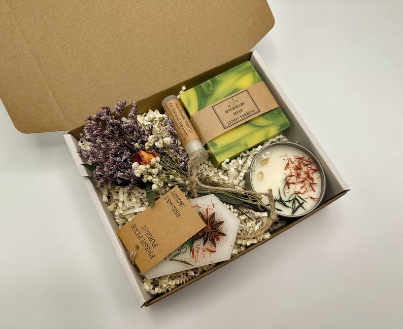 May include: A gift box filled with a green bar of soap labelled "Handmade Soap Lemon Verbena", a small white candle with red and yellow flower petals, a small tube of lip balm, and a small tag with the text "Fresh Pine,  Potpourri,  Shower,  Rosemary".