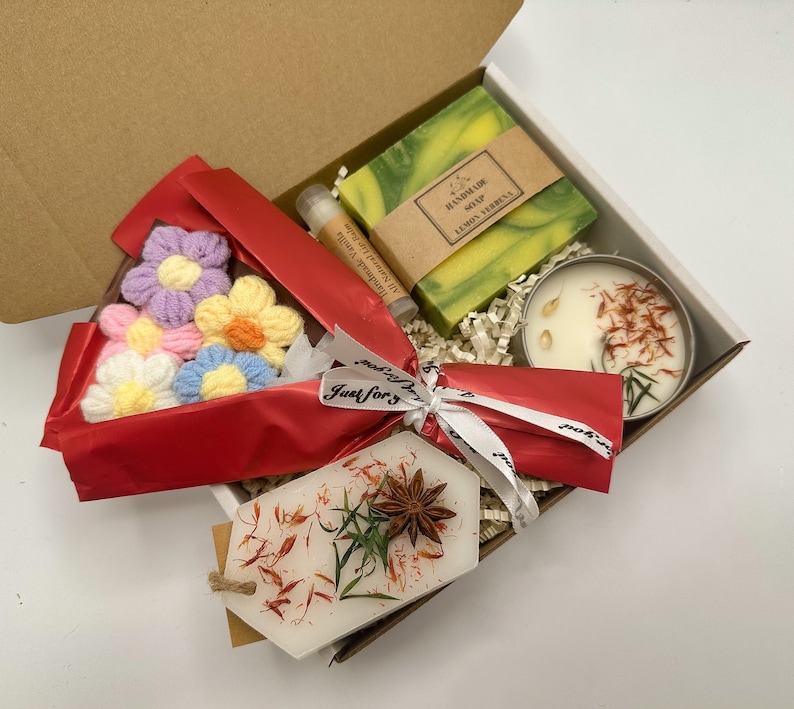 May include: A gift box filled with a variety of items, including a handmade soap bar with the text "Handmade Soap Lemon Verbena", a candle with dried flowers, a small bouquet of crocheted flowers, and a small tag that says "Just for you".