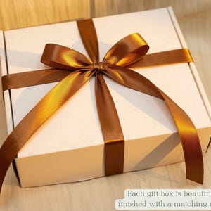 May include: A square white gift box tied with a shiny bronze-coloured satin ribbon arranged in a bow on top. The box features a small white label in the bottom right corner with the text: Each gift box is beautifully finished with a matching ribbon.
