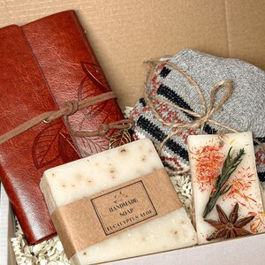 May include: A gift box filled with a brown leather journal, a white bar of handmade eucalyptus soap, a gray and brown knitted scarf, and a scented candle with dried flowers and star anise.