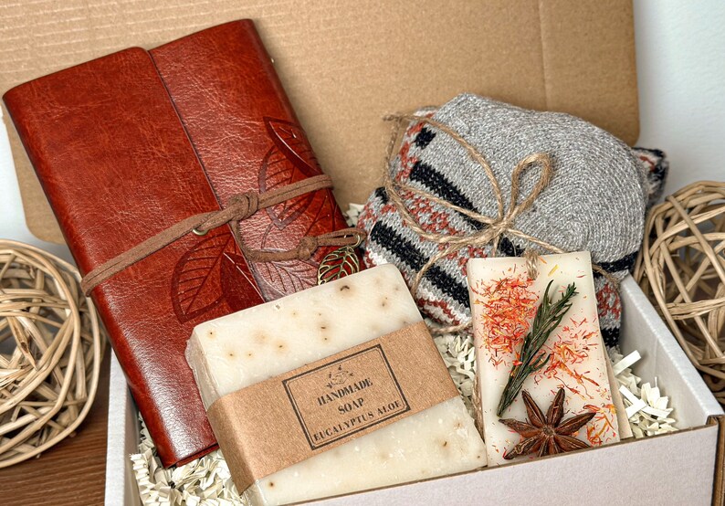 May include: A gift box filled with a brown leather journal, a white bar of handmade eucalyptus soap, a gray and brown knitted scarf, and a scented candle with dried flowers and star anise.