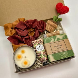 May include: A gift box containing two fabric scrunchies in mustard and maroon, a crocheted heart plant, a tube of shea butter hand and body cream, a bar of handmade English Garden soap, and a candle in a tin with three daisy-shaped wax embeds.