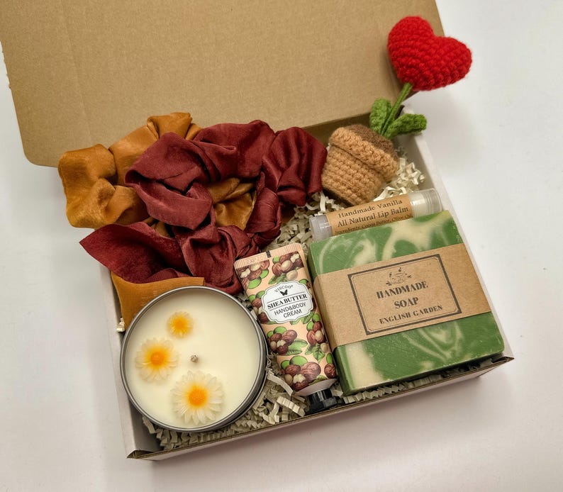 May include: A gift box containing two fabric scrunchies in mustard and maroon, a crocheted heart plant, a tube of shea butter hand and body cream, a bar of handmade English Garden soap, and a candle in a tin with three daisy-shaped wax embeds.