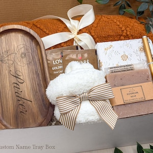May include: A gift box containing an oval wooden tray engraved with the name Parker and a floral design, a burnt orange knit blanket with a white ribbon, a white notebook, a gold pen, a lip balm, and a bar of eucalyptus aloe soap.