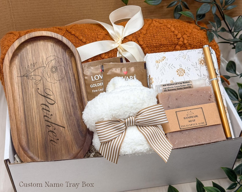 May include: A gift box containing an oval wooden tray engraved with the name Parker and a floral design, a burnt orange knit blanket with a white ribbon, a white notebook, a gold pen, a lip balm, and a bar of eucalyptus aloe soap.