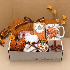 May include: A gift box labelled Autumn Warmth Box containing a textured orange blanket, a Ferrero Rocher chocolate bar, a white mug with pumpkin and leaf illustrations, a ceramic pumpkin, a white towel, a candle, and a wrapped soap bar.