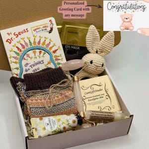 May include: A gift box containing a Dr. Seuss book, a crocheted bunny, socks, a rose quartz face roller, eye masks, chamomile hand cream, soap, and lip balm. A greeting card with the word "Congratulations" is also included.