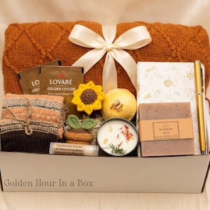 May include: A gift box labeled Golden Hour In a Box containing a rust-colored knit blanket with a white ribbon, Lovar&eacute; tea, a crochet sunflower, a bath bomb, a floral notebook, a gold pen, eucalyptus aloe soap, a candle, and vanilla lip balm.