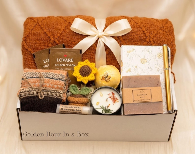 May include: A gift box labeled Golden Hour In a Box containing a rust-colored knit blanket with a white ribbon, Lovar&eacute; tea, a crochet sunflower, a bath bomb, a floral notebook, a gold pen, eucalyptus aloe soap, a candle, and vanilla lip balm.
