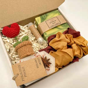 May include: A gift box filled with a variety of items, including a crocheted flower, a handmade soap bar, a lip balm, a satin scrunchie, and a small brown paper bag with the text "Fresh Pine Forest Beeswax Sachet".