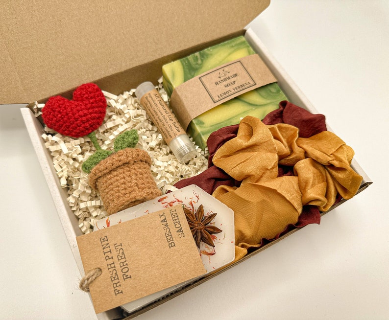 May include: A gift box filled with a variety of items, including a crocheted flower, a handmade soap bar, a lip balm, a satin scrunchie, and a small brown paper bag with the text "Fresh Pine Forest Beeswax Sachet".