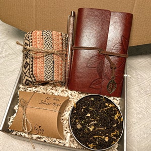 May include: A gift box filled with a brown leather journal with a leaf design, a pair of orange and brown patterned socks, a small brown paper bag with 10 tea infuser bags, and a round tin of loose leaf tea.