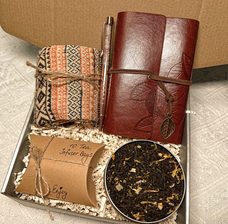 May include: A gift box filled with a brown leather journal with a leaf design, a pair of orange and brown patterned socks, a small brown paper bag with 10 tea infuser bags, and a round tin of loose leaf tea.