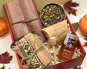 May include: A red gift box filled with a brown leather journal, a pair of knitted socks, a jar of honey, a small bag of tea, and a wooden honey dipper. The box is decorated with autumn leaves and a small pine cone.