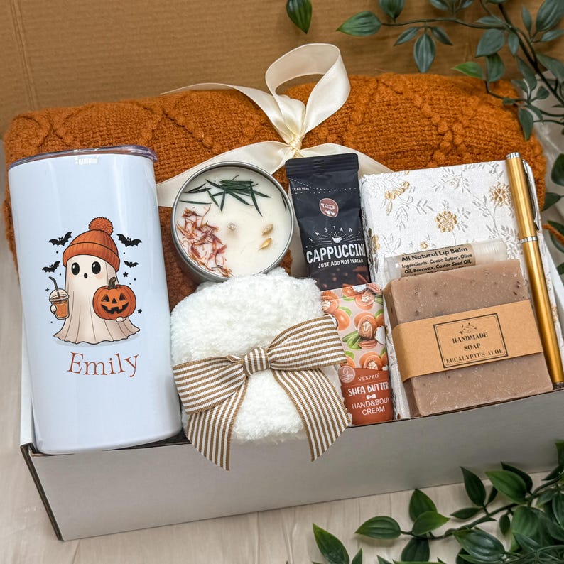 May include: A gift box containing a white tumbler with a ghost graphic and the name Emily, a rust-coloured knit blanket, a candle, a cappuccino packet, a white fuzzy item with a striped bow, a floral notebook, a gold pen, lip balm, and soap.