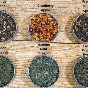 Op de afbeelding: Zes ronde metalen blikken gevuld met losse bladthee. De blikken zijn gerangschikt in twee rijen van drie. De bovenste rij van links naar rechts is gelabeld "Candy Cane Burst", "Cranberry Apple" en "Frosty Plum Spice". De onderste rij van links naar rechts is gelabeld "Green Peacock", "Jasmine Gold Dragon" en "Lucky Dragon Hyson".