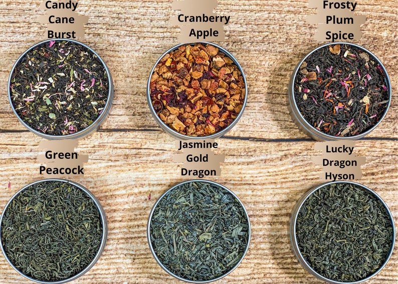 Op de afbeelding: Zes ronde metalen blikken gevuld met losse bladthee. De blikken zijn gerangschikt in twee rijen van drie. De bovenste rij van links naar rechts is gelabeld "Candy Cane Burst", "Cranberry Apple" en "Frosty Plum Spice". De onderste rij van links naar rechts is gelabeld "Green Peacock", "Jasmine Gold Dragon" en "Lucky Dragon Hyson".