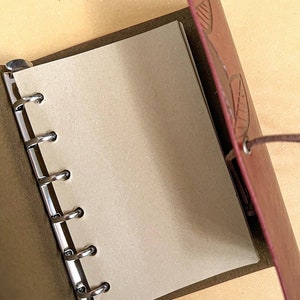 May include: A brown leather bound journal with a leaf design embossed on the cover. The journal has a metal ring binding system and is open to a blank page.
