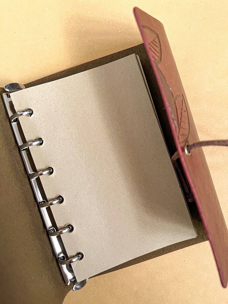 May include: A brown leather bound journal with a leaf design embossed on the cover. The journal has a metal ring binding system and is open to a blank page.