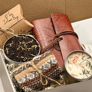 May include: A gift box filled with a brown leather journal, a tin of loose leaf tea, a pair of knitted socks, and a candle with dried herbs and spices. The box is lined with brown paper shreds.