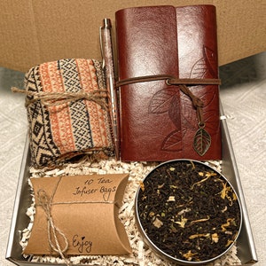 May include: A gift box filled with a brown leather journal, a pen, a pair of socks, a small bag of tea, and a brown paper bag with the text "10 Tea Infuser Bags" and "Enjoy".