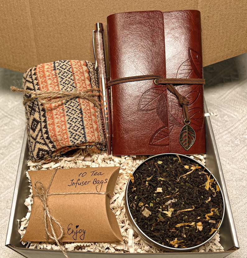 May include: A gift box filled with a brown leather journal, a pen, a pair of socks, a small bag of tea, and a brown paper bag with the text "10 Tea Infuser Bags" and "Enjoy".