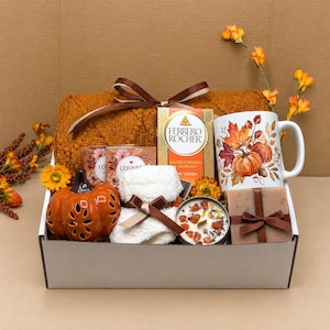 May include: A gift box containing a textured orange blanket, a white mug with pumpkin and leaf illustrations, a box of Ferrero Rocher chocolates, a ceramic orange pumpkin, a white fuzzy cloth, a candle with dried toppings, and a wrapped soap bar.