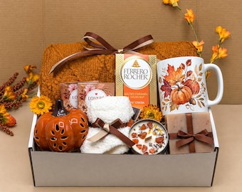Op de afbeelding: Een geschenkdoos met een oranje deken met textuur, een witte mok met pompoen- en bladafbeeldingen, een doos Ferrero Rocher chocolaatjes, een oranje keramische pompoen, een witte pluizige doek, een kaars met decoratie en een verpakt stuk zeep.