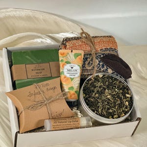 May include: A gift box containing a green bar of handmade soap labeled English Garden, a tube of melon hand and body cream, a pair of patterned knit socks, a tin of loose leaf tea, a brown paper pillow box labeled Tea Infuser Bags, and lip balm.