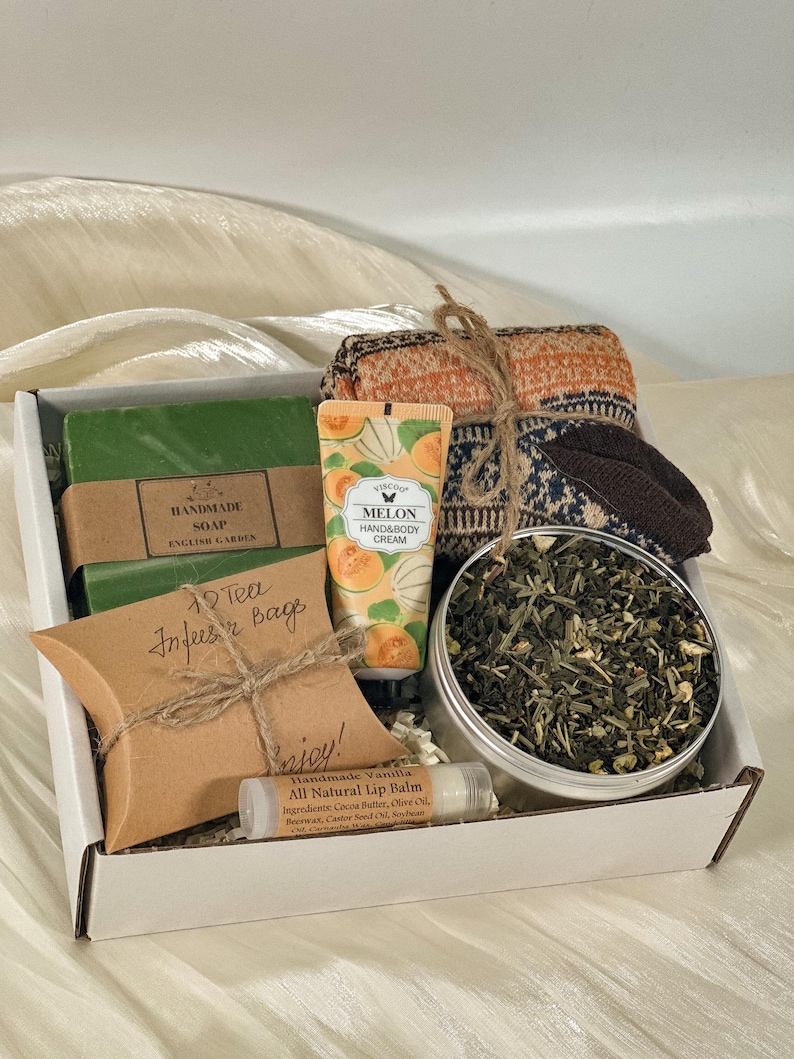 May include: A gift box containing a green bar of handmade soap labeled English Garden, a tube of melon hand and body cream, a pair of patterned knit socks, a tin of loose leaf tea, a brown paper pillow box labeled Tea Infuser Bags, and lip balm.