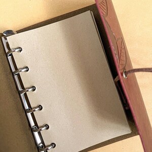 May include: A brown leather bound journal with a leaf design embossed on the cover. The journal has a metal ring binding system and is open to a blank page.
