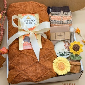 May include: A gift box containing a textured rust-colored blanket tied with a white ribbon, a Ferrero Rocher chocolate bar, patterned socks, a bar of handmade soap, a lip balm, a round candle, and a small crocheted sunflower in a pot.