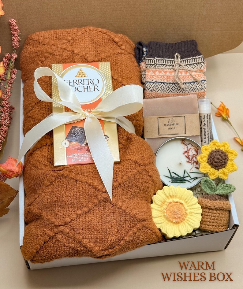 May include: A gift box containing a textured rust-colored blanket tied with a white ribbon, a Ferrero Rocher chocolate bar, patterned socks, a bar of handmade soap, a lip balm, a round candle, and a small crocheted sunflower in a pot.