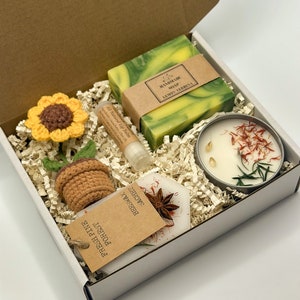 May include: A white gift box filled with a variety of items, including a green and yellow bar of soap labelled "Handmade Soap Lemon Verbena", a small jar of candle with dried flowers and herbs, a lip balm tube, a crocheted sunflower, a crocheted pot, and a small brown paper bag with the text "Forest Fresh Pine Sachet Beeswax".