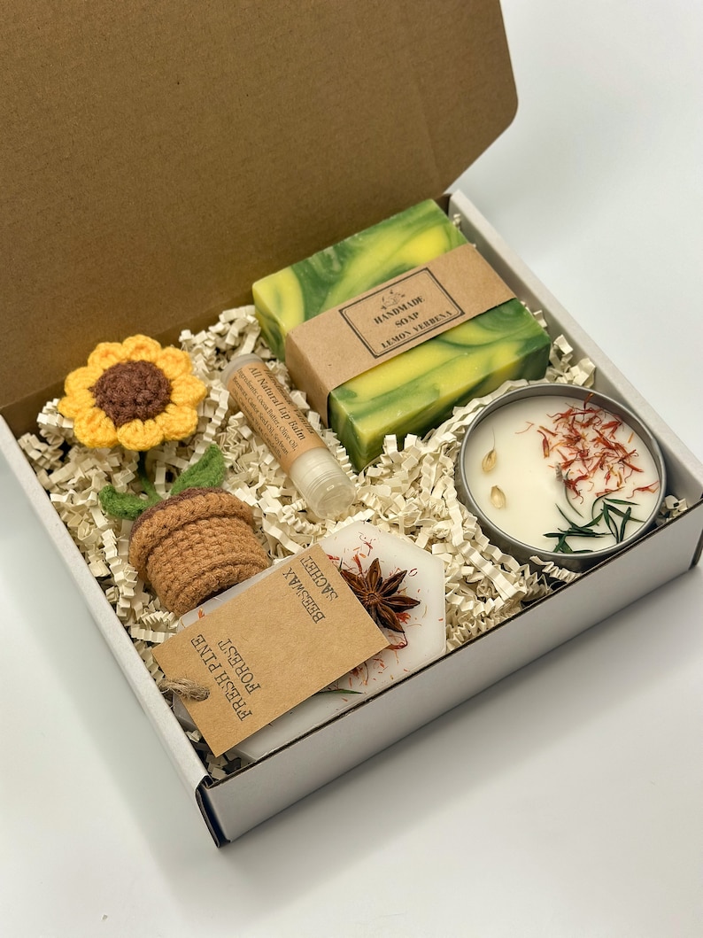 May include: A white gift box filled with a variety of items, including a green and yellow bar of soap labelled "Handmade Soap Lemon Verbena", a small jar of candle with dried flowers and herbs, a lip balm tube, a crocheted sunflower, a crocheted pot, and a small brown paper bag with the text "Forest Fresh Pine Sachet Beeswax".