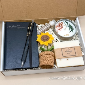 May include: A gift box containing a black notebook with the name "Andrea", a pen, a lip balm, a crochet sunflower in a pot, a candle, and a bar of handmade soap. The box is labeled "Custom Notebook Gift Box".