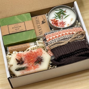 May include: A gift box containing a green bar of handmade soap labeled English Garden, a box of matches, a round candle with botanical toppings, a tube of vanilla lip balm, a white wax melt with star anise, and a pair of brown patterned socks.