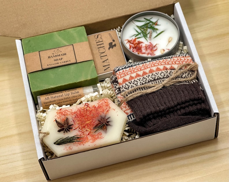 May include: A gift box containing a green bar of handmade soap labeled English Garden, a box of matches, a round candle with botanical toppings, a tube of vanilla lip balm, a white wax melt with star anise, and a pair of brown patterned socks.