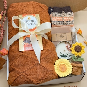 May include: A gift box containing a rust-coloured textured blanket tied with a white ribbon, a Ferrero Rocher chocolate bar, patterned knit socks, a bar of handmade soap, a lip balm, a round candle, and a crocheted sunflower in a pot.
