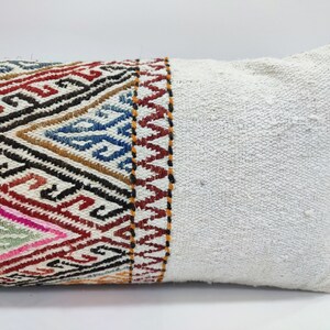 May include: A white lumbar pillow with a colourful geometric patterned band along one side. The pillow is made of a soft, woven fabric.