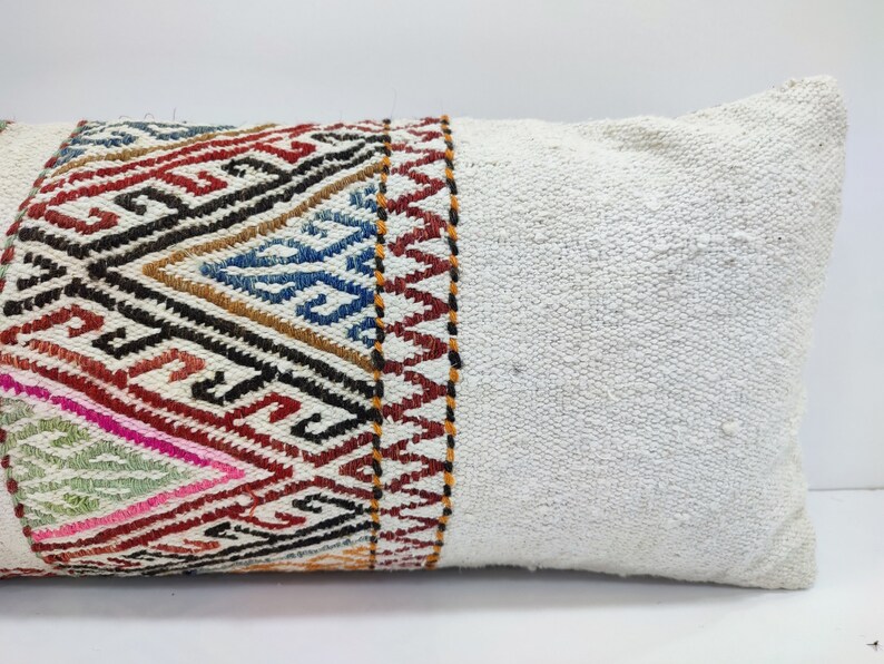 May include: A white lumbar pillow with a colourful geometric patterned band along one side. The pillow is made of a soft, woven fabric.