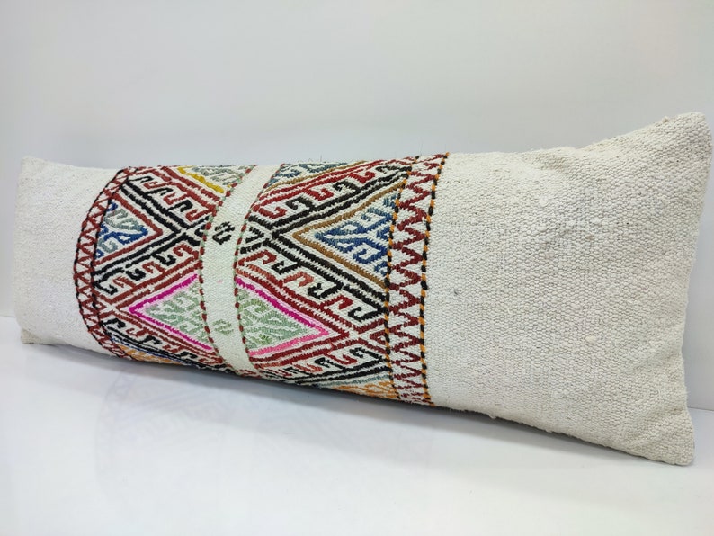 May include: A white lumbar pillow with a colourful geometric pattern woven into the fabric. The pattern is a mix of red, blue, green, yellow, and black.