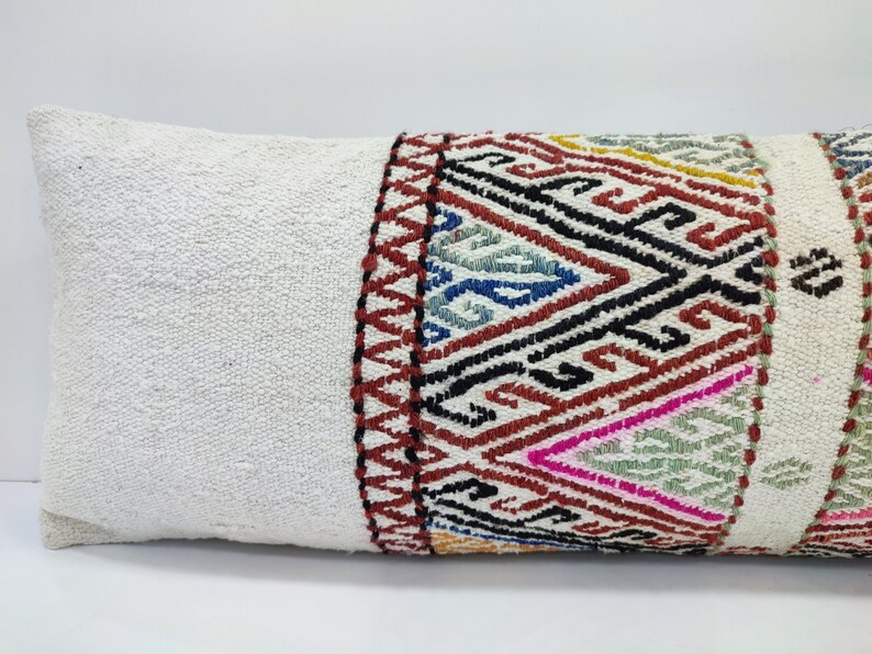 May include: White pillow with a colourful geometric pattern on one end. The pattern is made up of red, black, blue, green, and yellow threads.