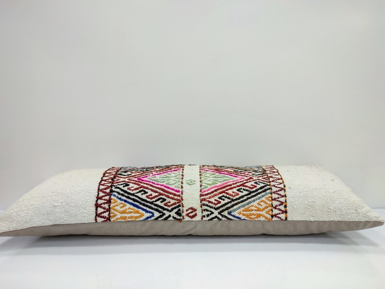 May include: A long, rectangular white cushion with a colourful geometric pattern woven into the centre. The cushion has a beige backing.