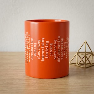 May include: A bright orange ceramic mug featuring a vertical list of white text including words like crystallizing, razzle-dazzling, accomplishing, ruminating, musing, sleuthing, improvising, ionizing, jiving, actioning, deliberating, cogitating, baking, and frolicking.
