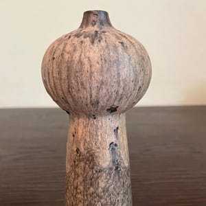 May include: A small, handcrafted ceramic vase with a bulbous top and a slender, textured base. The vase features a mottled, earthy color palette of brown, tan, and dark gray, creating a rustic aesthetic. The vase is approximately 6 inches tall.