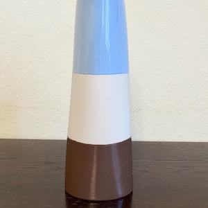 Könnte beinhalten: Eine hohe Keramikvase mit konischer Form. Die Vase ist in drei horizontale Abschnitte unterteilt: hellblau oben, weiß in der Mitte und braun unten. Die Vase steht auf einer dunklen Holzoberfläche.