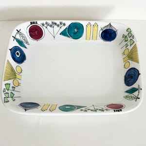 Può includere: Un piatto da portata rettangolare in ceramica bianca con un bordo decorativo di illustrazioni dipinte a mano. Il design include elementi botanici stilizzati, forme astratte e motivi circolari in tonalità blu, turchese, giallo, verde e rosso.